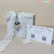 A Pearl Lace Bow Keychain with Silver Heart – Elegant Bag Charm from Tiny Mochi sits beside a brand business card with QR codes and social info, all arranged on a light beige surface for a coquette-inspired display.