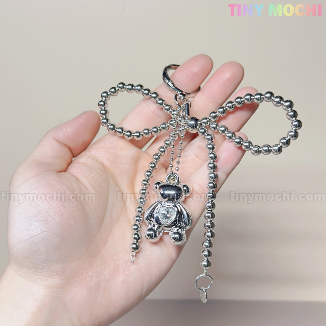 A hand displays the Silver Teddy Bear Bow Keychain by Tiny Mochi, a cute silver bow-shaped bag charm with a dangling teddy bear. Tiny Mochi appears in pastel at the top right on a neutral background—ideal for women, teens, and Kawaii lovers!.