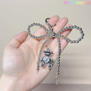 A hand displays the Silver Teddy Bear Bow Keychain by Tiny Mochi, a cute silver bow-shaped bag charm with a dangling teddy bear. Tiny Mochi appears in pastel at the top right on a neutral background—ideal for women, teens, and Kawaii lovers!.
