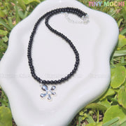 The Black Beaded Flower Necklace with Silver Floral Charm by Tiny Mochi, a handmade Y2K choker with a soft grunge aesthetic, is displayed on a wavy white dish atop green grass, with the Tiny Mochi logo in the top right corner.