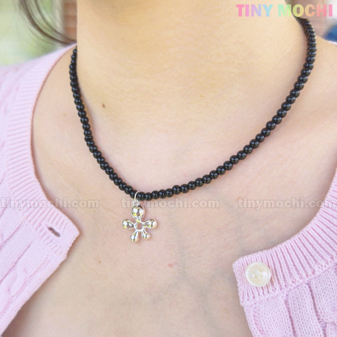 A person wears Tiny Mochis Black Beaded Flower Necklace with Silver Floral Charm, styled with a light pink buttoned sweater for a Y2K-inspired look. The TINY MOCHI logo appears in the top right corner of the image.