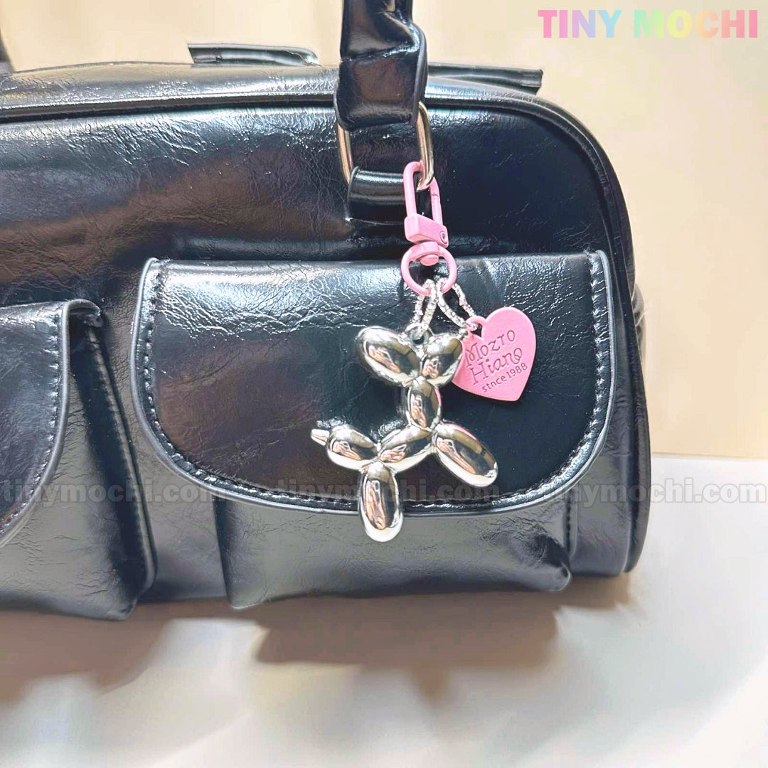 A shiny black handbag with front pockets exudes soft girl aesthetics, featuring Tiny Mochi’s Balloon Dog Keychain Chrome Silver Bag Charm with a pink heart tag for a playful touch.