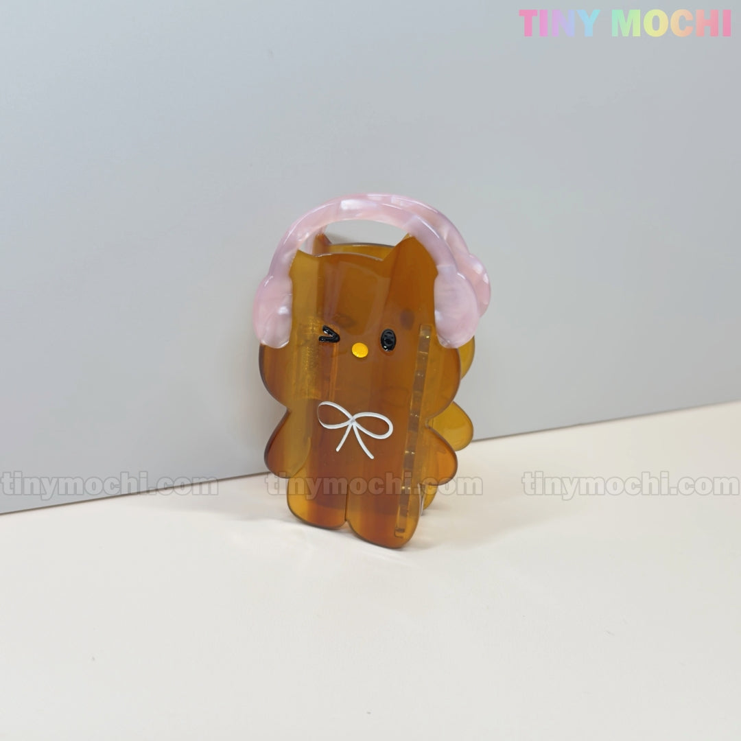 A cute brown bear-shaped Kawaii Bunny & Bear Hair Claw Clip with pink headphones and a white bow stands on a white surface; TINY MOCHI is displayed in pastel colors at the top right.