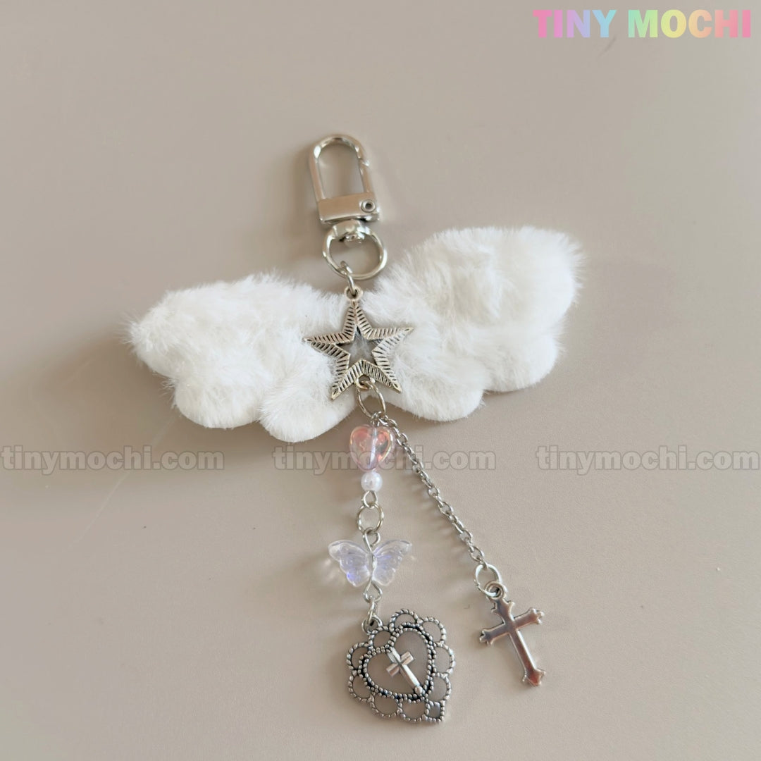 Tiny Mochi’s Fluffy Angel Wing Keychain – Star & Cross Bag Charm Clip features white angel wings, a plush angel, silver star, and dangling purple butterfly, heart with cross, and silver cross on a beige background.