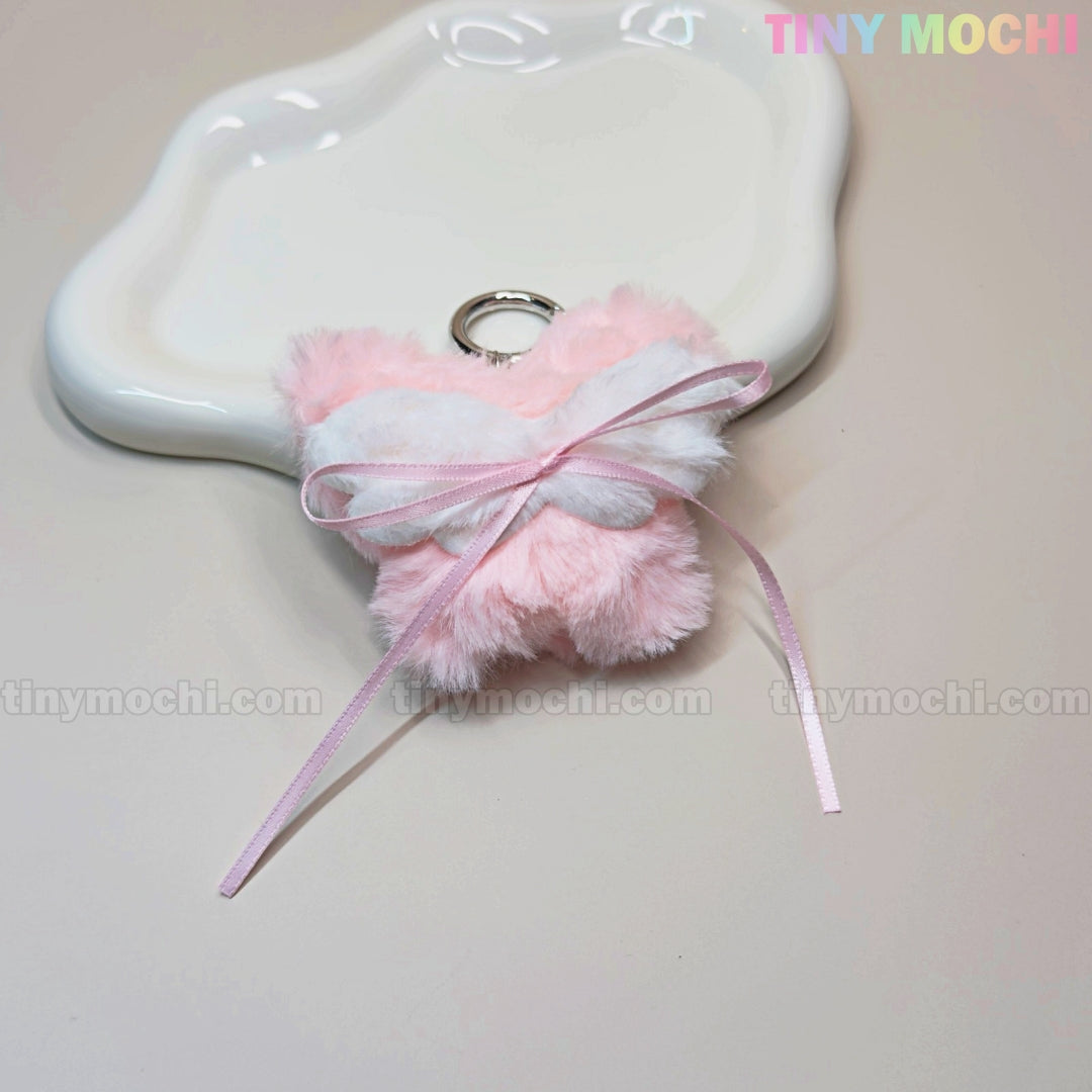 The Tiny Mochi Plush Butterfly Bow Keychain, a pink fuzzy bag charm with a white bow and ribbon for a coquette-inspired style, is displayed on a wavy tray with the TINY MOCHI logo at the top right.