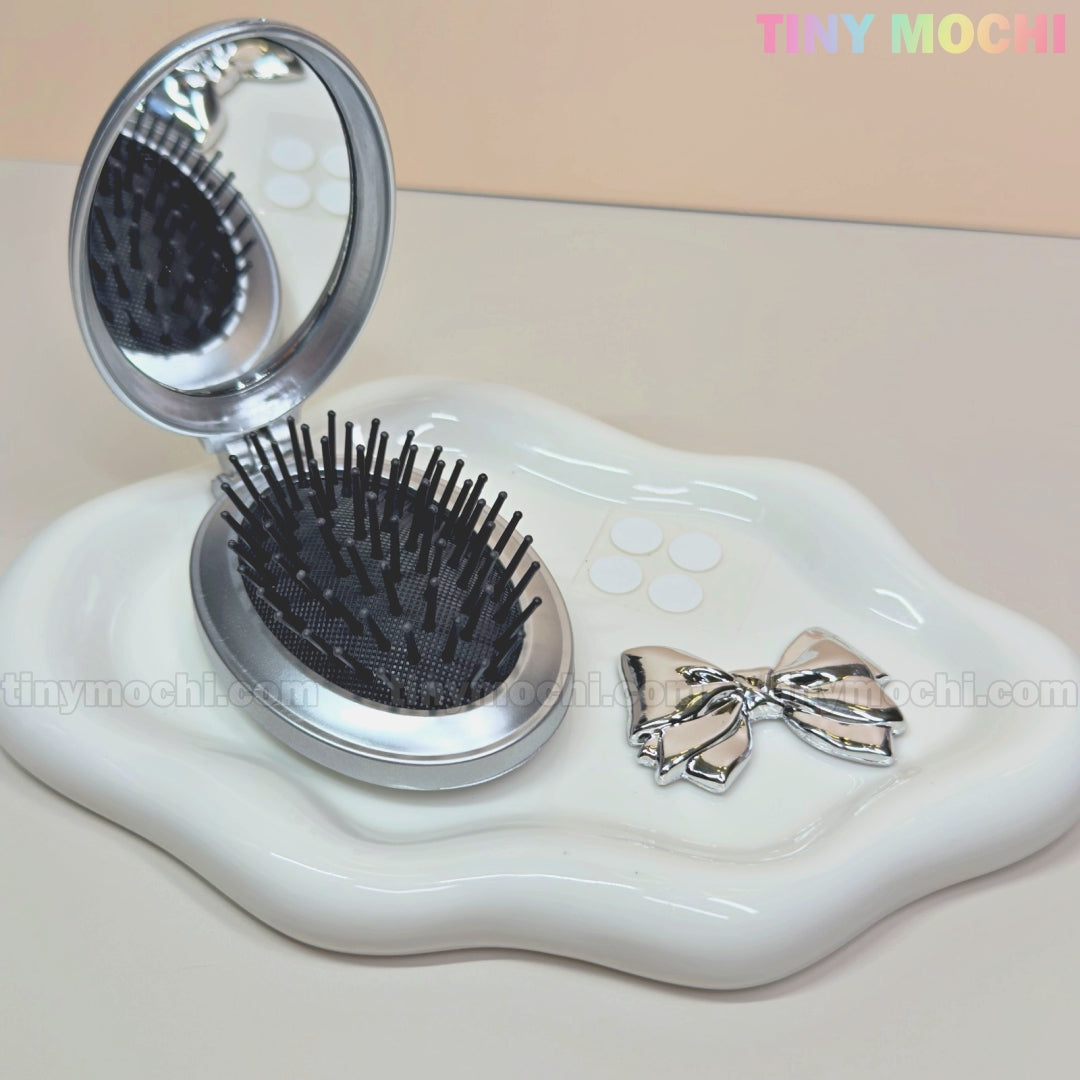 The Tiny Mochi Oval Silver Folding Travel Mirror Comb, a compact pocket-sized hairbrush with a built-in mirror, is displayed open on a wavy-edged tray next to a silver bow. The beige background displays TINY MOCHI in the corner.