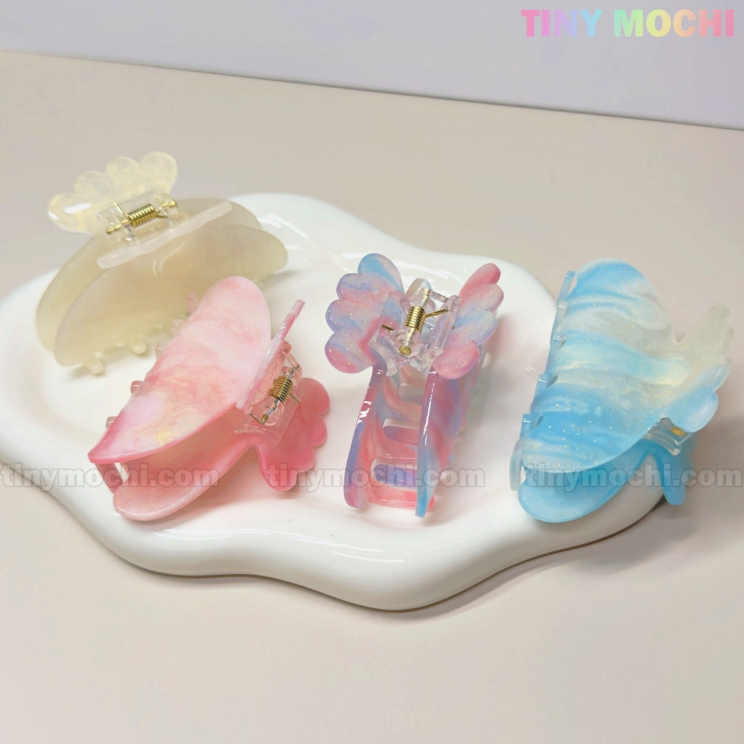 Three pastel butterfly-shaped UV reactive acetate hair claw clips from Tiny Mochi change color in sunlight. With a marble-like finish, these fairycore accessories in cream, pink, and blue add a magical touch to any look.