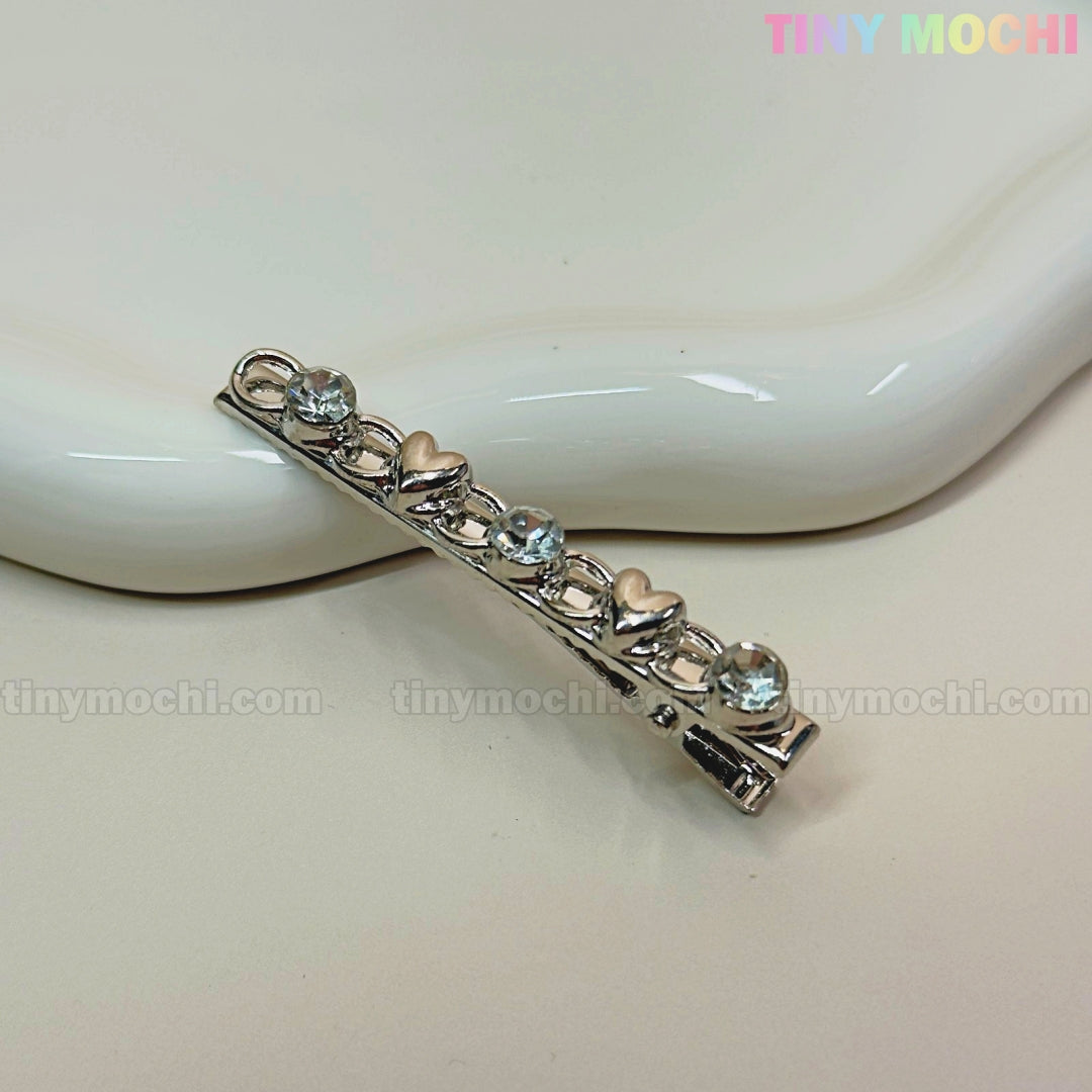 A silver Kawaii Gem Hair Clip for Girls by Tiny Mochi, decorated with heart shapes and clear gemstones, rests on white ceramic. The plain background shows Tiny Mochi in pastel colors in the top right corner. Perfect Y2K coquette accessory!.