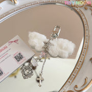 The Fluffy Angel Wing Keychain – Star & Cross Bag Charm Clip by Tiny Mochi features white plush wings and silver star, cross, butterfly, and heart charms, exuding a gothic angel vibe as it rests on an ornate mirror next to a Tiny Mochi card.