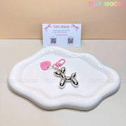 A Tiny Mochi Balloon Dog Keychain Chrome Silver Bag Charm with a pink heart tag sits on a wavy white dish, while a Tiny Mochi business card in the background enhances the soft girl, Y2K-inspired aesthetic.