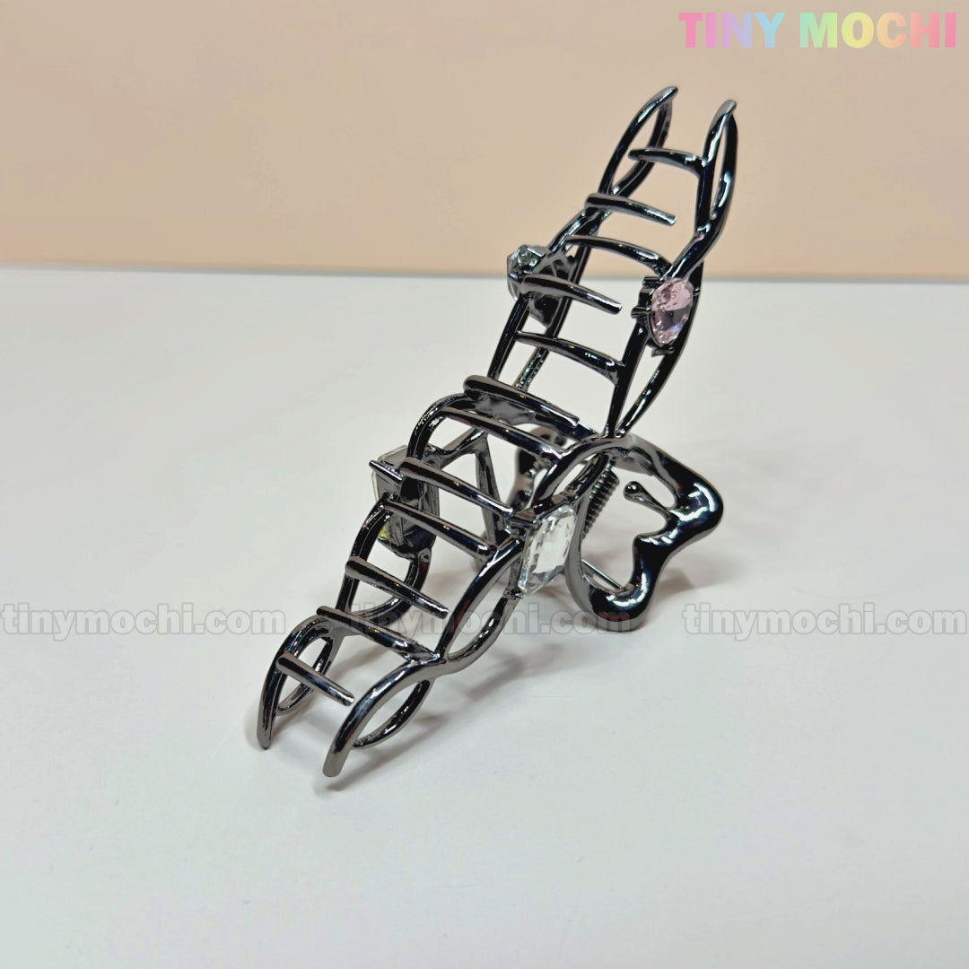 The Dark Silver Crystal Heart Hair Claw by Tiny Mochi, an elegant metal hair clip adorned with crystals, is displayed on a white surface. The brand name, Tiny Mochi, appears in pastel colors at the top right.