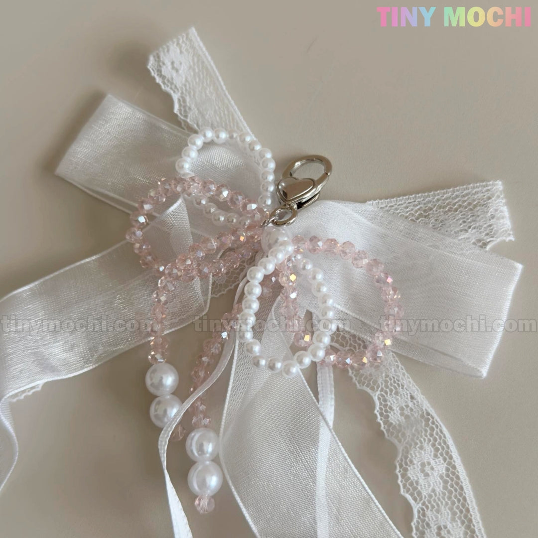 The Lace Bow Ribbon Keychain – Pearl Angelcore Bag Charm Clip by Tiny Mochi features white lace, sheer ribbon, pink and white pearl beads in loops, a silver clasp at the center, and the Tiny Mochi logo in the top right corner.