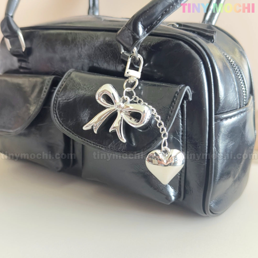 A shiny black handbag with a front pocket is displayed, featuring Tiny Mochi’s Silver Bow Heart Keychain – a kawaii mirror bag charm for backpacks. The light background highlights the coquette style, with the Tiny Mochi logo in the top right corner.