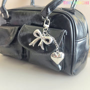 A shiny black handbag with a front pocket is displayed, featuring Tiny Mochi’s Silver Bow Heart Keychain – a kawaii mirror bag charm for backpacks. The light background highlights the coquette style, with the Tiny Mochi logo in the top right corner.