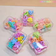 Five Tiny Mochi Kids Hair Clips Sets, each in a clear mini travel case, featuring 10 colorful kawaii hair claws in butterfly, flower, heart and more fun designs, arranged in a circle on a light surface.