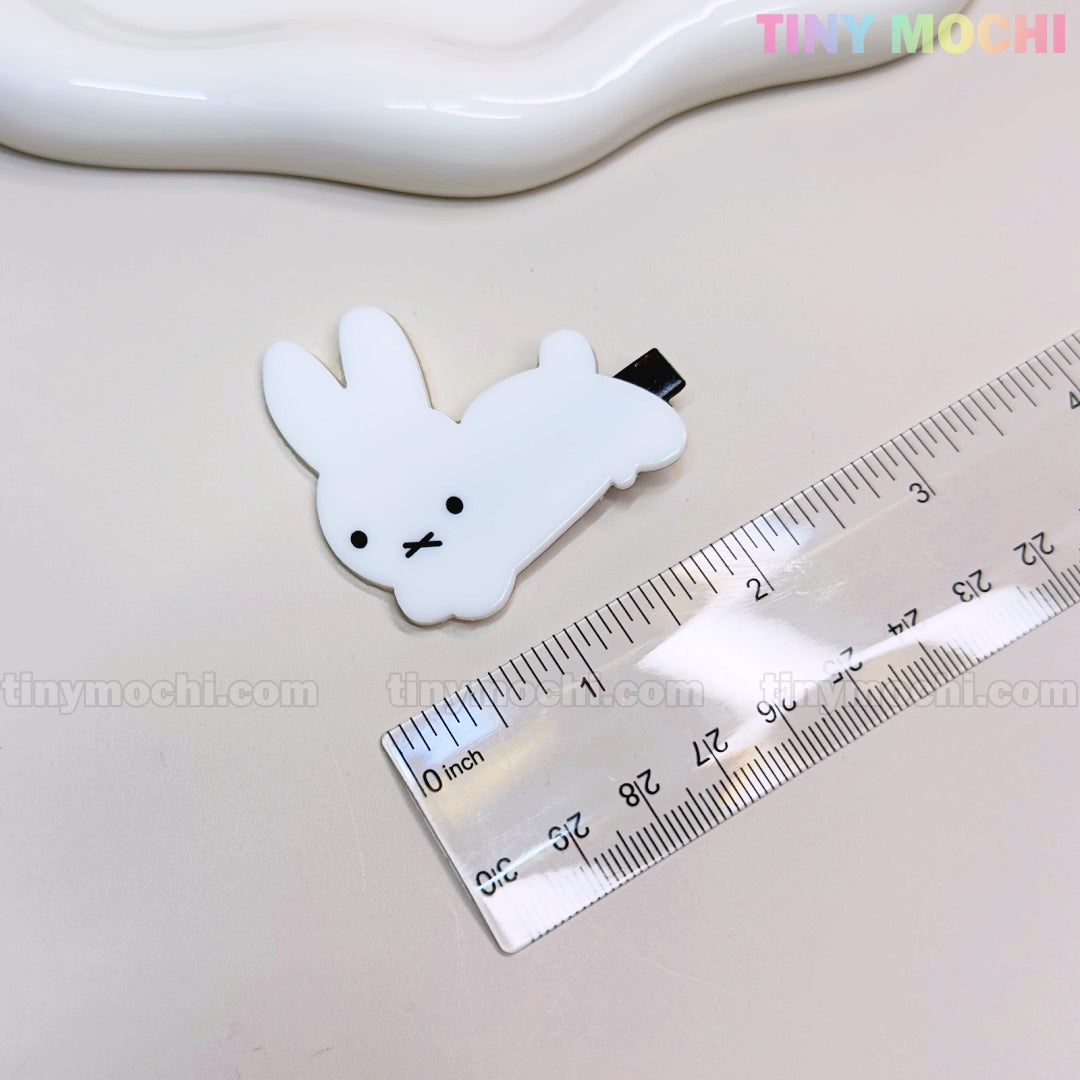 A small, white bunny from the “Kawaii Bunny and Bear Hair Clips for Girls” by Tiny Mochi rests next to a ruler on a beige surface. The clip features black eyes, an “X” mouth, and the Tiny Mochi logo appears in the top right corner.