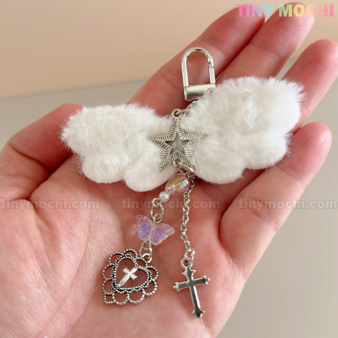 A hand displays the Fluffy Angel Wing Keychain – Star & Cross Bag Charm Clip by Tiny Mochi, featuring white wings with silver star, cross, heart, and purple butterfly charms against a neutral background; “Tiny Mochi” appears in the corner.