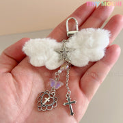 A hand displays the Fluffy Angel Wing Keychain – Star & Cross Bag Charm Clip by Tiny Mochi, featuring white wings with silver star, cross, heart, and purple butterfly charms against a neutral background; “Tiny Mochi” appears in the corner.