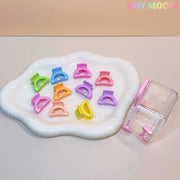 A white, irregular dish displays the Tiny Mochi Kids Hair Clips Set with 10 colorful kawaii butterfly, flower, and heart hair claws. Beside it is a clear mini travel case with a pink handle and one clip inside.