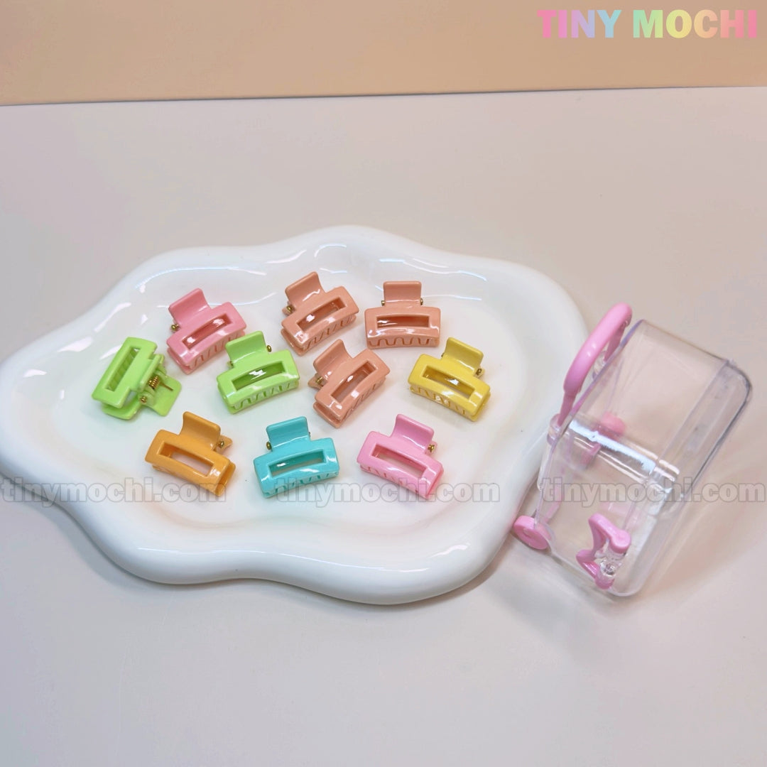 A wavy white dish displays Tiny Mochi’s Kids Hair Clips Set: 10 colorful kawaii mini claws in butterfly, flower, and heart shapes. Beside it is a clear suitcase with a pink handle for storing these cute accessories on the go.