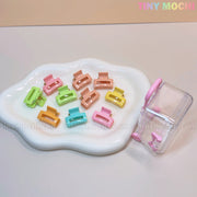 A wavy white dish displays Tiny Mochi’s Kids Hair Clips Set: 10 colorful kawaii mini claws in butterfly, flower, and heart shapes. Beside it is a clear suitcase with a pink handle for storing these cute accessories on the go.