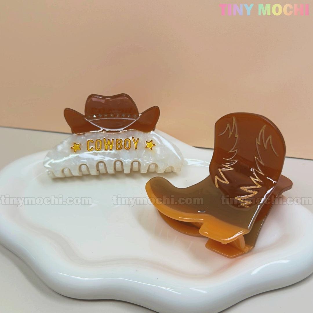 Two Western Cowboy Hair Claw Clips by Tiny Mochi—a brown hat with “COWBOY” and a brown boot—sit on a wavy white tray. These cute cowgirl barrettes are perfect western hair accessories for women and girls. Pastel Tiny Mochi branding appears in the top right.