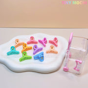 A white tray displays the Tiny Mochi Kids Hair Clips Set—10 colorful kawaii mini hair claws (butterfly, flower & heart designs). Beside it is a clear travel case with pink clasp and handle, all set on a light apricot background.