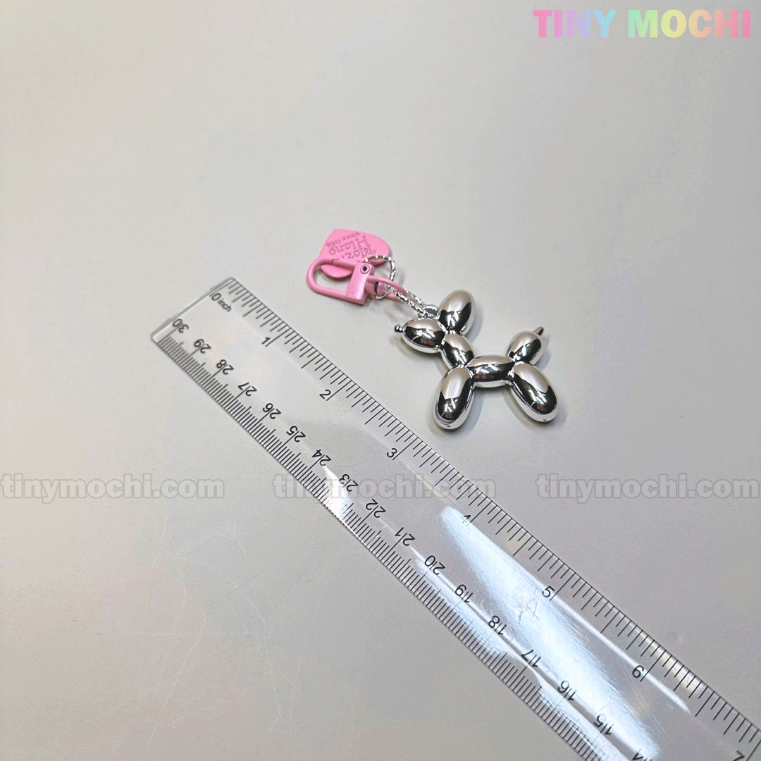 The Balloon Dog Keychain Chrome Silver Bag Charm with Pink Heart Tag from Tiny Mochi shines next to a clear ruler on a light surface, adding Y2K-inspired, soft girl flair. tinymochi.com appears subtly in the background.
