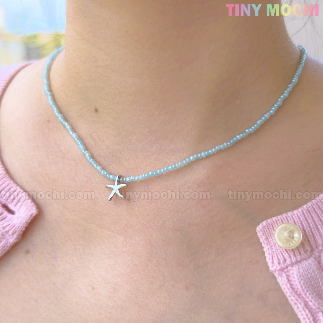 A close-up shows a person wearing the Tiny Mochi Silver Starfish Choker Necklace, a light blue beaded beach jewelry piece, paired with a pink sweater with a white button for oceancore style.
