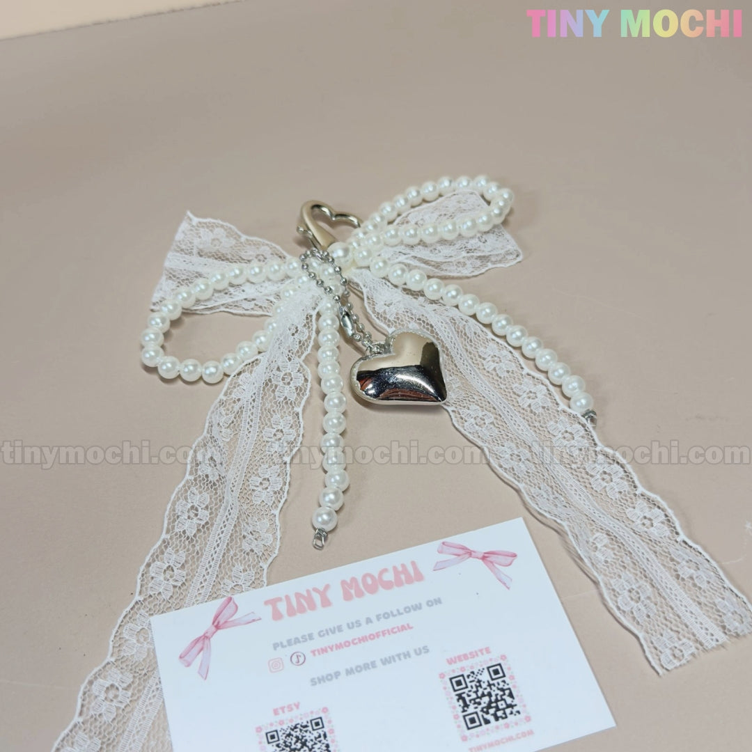 The Pearl Lace Bow Keychain with Silver Heart by Tiny Mochi features elegant white lace, faux pearls, and a silver heart charm—perfect as a chic bag accessory. Shown beside a Tiny Mochi business card on beige with the logo in the top right corner.