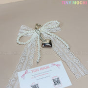 The Pearl Lace Bow Keychain with Silver Heart by Tiny Mochi features elegant white lace, faux pearls, and a silver heart charm—perfect as a chic bag accessory. Shown beside a Tiny Mochi business card on beige with the logo in the top right corner.