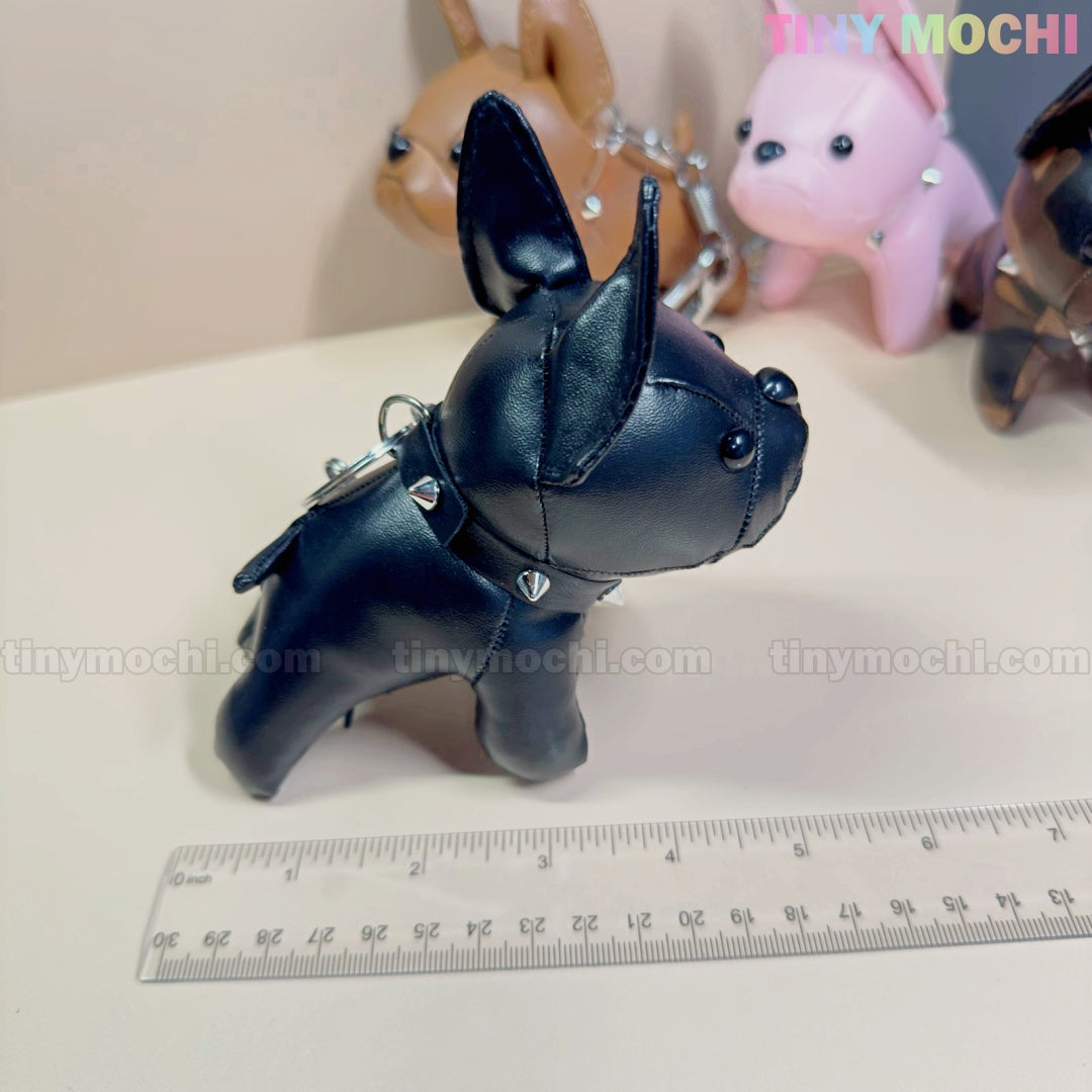 The Tiny Mochi French Bulldog Keychain Coin Purse, a mini faux leather dog bag charm with a studded collar, stands by a ruler—brown, pink, and black puppy coin purses blurred in the background. Perfect zipper pouch gift for dog lovers.