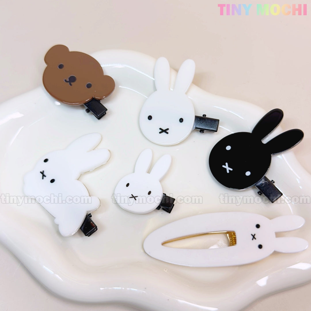 A white plate holds six Tiny Mochi Kawaii Bunny and Bear Hair Clips for Girls—cute cartoon animal snap and alligator clips featuring brown and black bears and white bunnies with minimalistic faces. Brand logo is top right.
