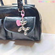 A sleek black handbag features a chrome silver Balloon Dog Keychain bag charm with a pink heart tag, adding cute Y2K flair. The brand name Tiny Mochi is displayed in pastel colors in the top right corner.