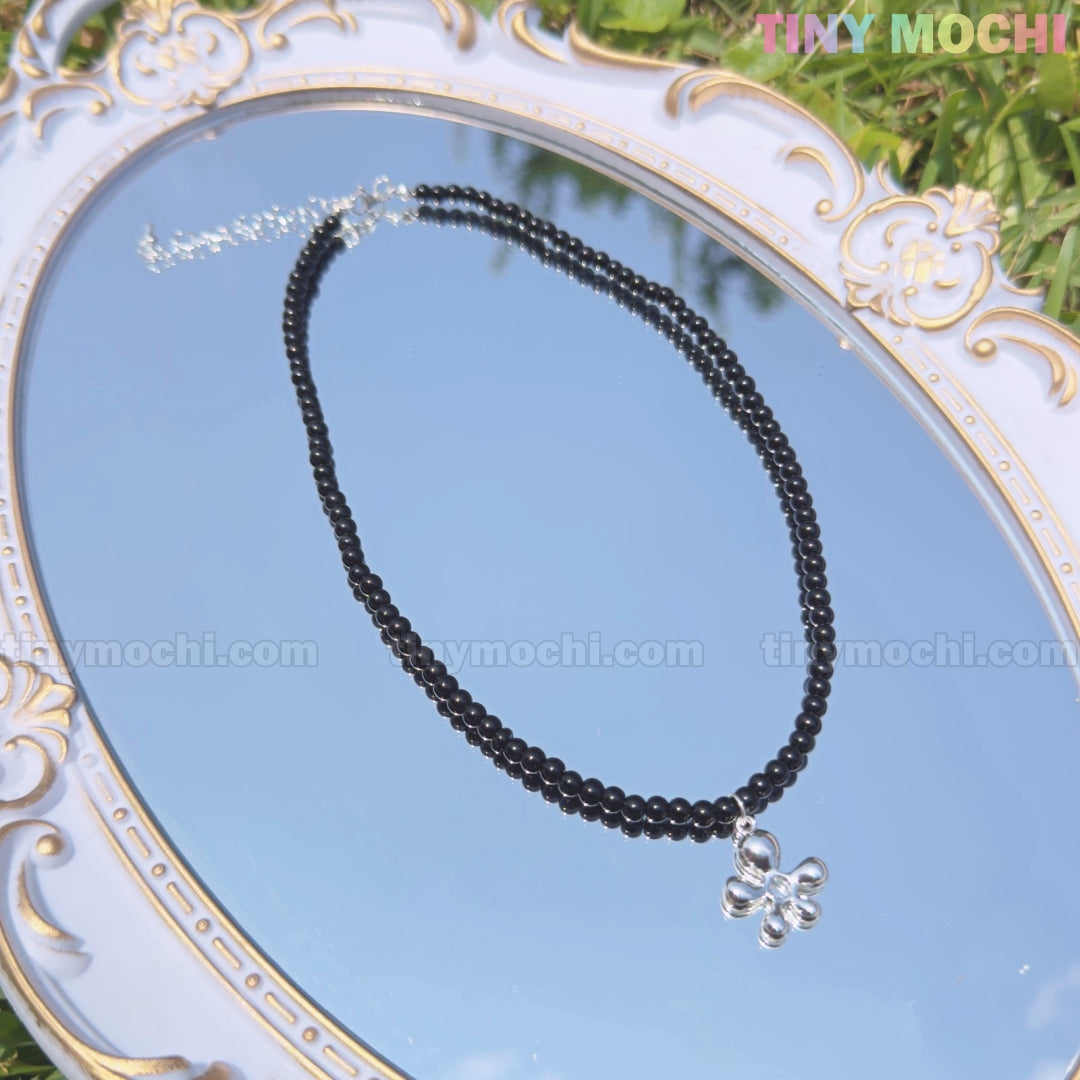 The Black Beaded Flower Necklace with Silver Floral Charm by Tiny Mochi rests on an ornate mirror atop grass, blending Y2K and soft grunge aesthetics. The sky reflects in the mirror, with Tiny Mochi branding in the corners.
