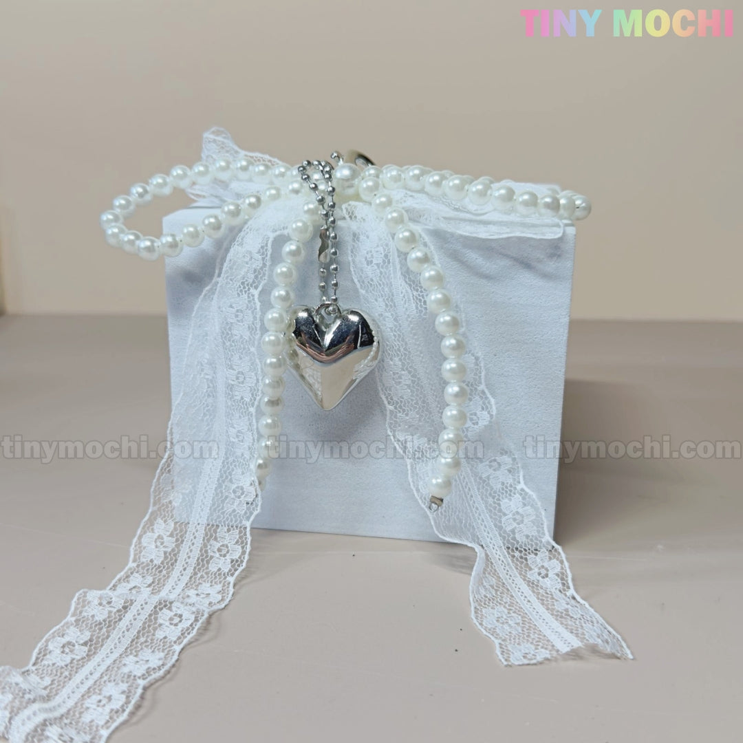 A white gift box with lace ribbon, faux pearls, and a silver heart channels coquette vibes. Against a light beige background, Tiny Mochi appears in pastels—ideal for the Pearl Lace Bow Keychain with Silver Heart, an elegant bag charm.