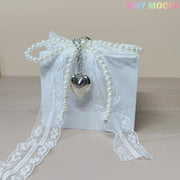 A white gift box with lace ribbon, faux pearls, and a silver heart channels coquette vibes. Against a light beige background, Tiny Mochi appears in pastels—ideal for the Pearl Lace Bow Keychain with Silver Heart, an elegant bag charm.
