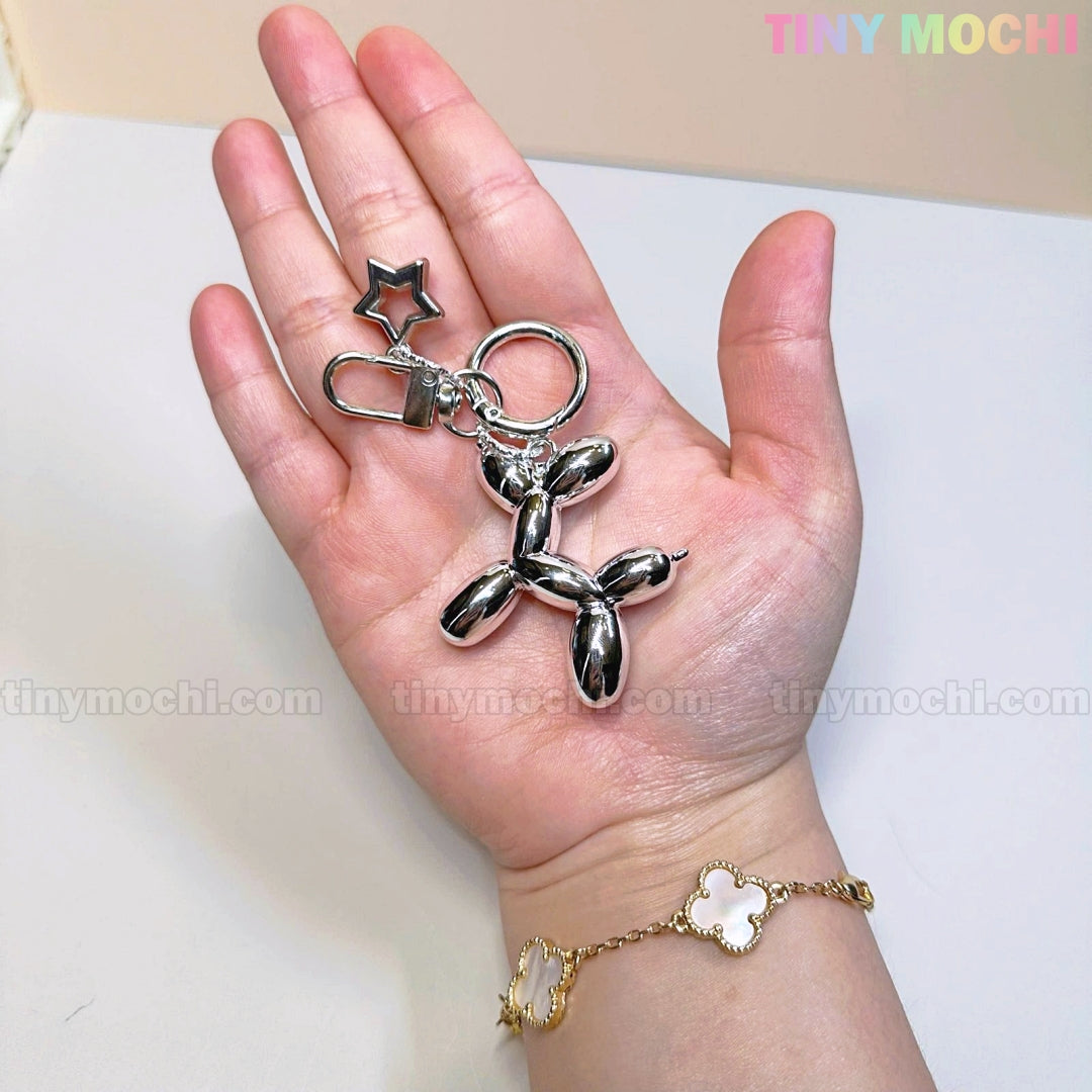 A hand holds the Tiny Mochi Balloon Dog Keychain Silver Chrome Bag Charm with a star accent. The person wears a gold bracelet with white flower charms. TINY MOCHI appears in pastel colors in the top right corner.