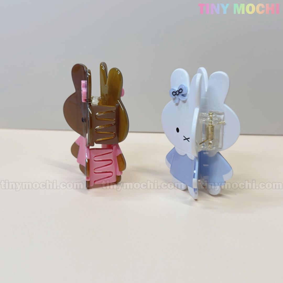 Two Kawaii Bunny Hair Claw Clips from Tiny Mochi stand upright: one brown with a pink dress, the other white with a blue dress and bow. These cute pastel acrylic bunny clips make perfect hair accessories. Tiny Mochi is shown in pastel at the top right.