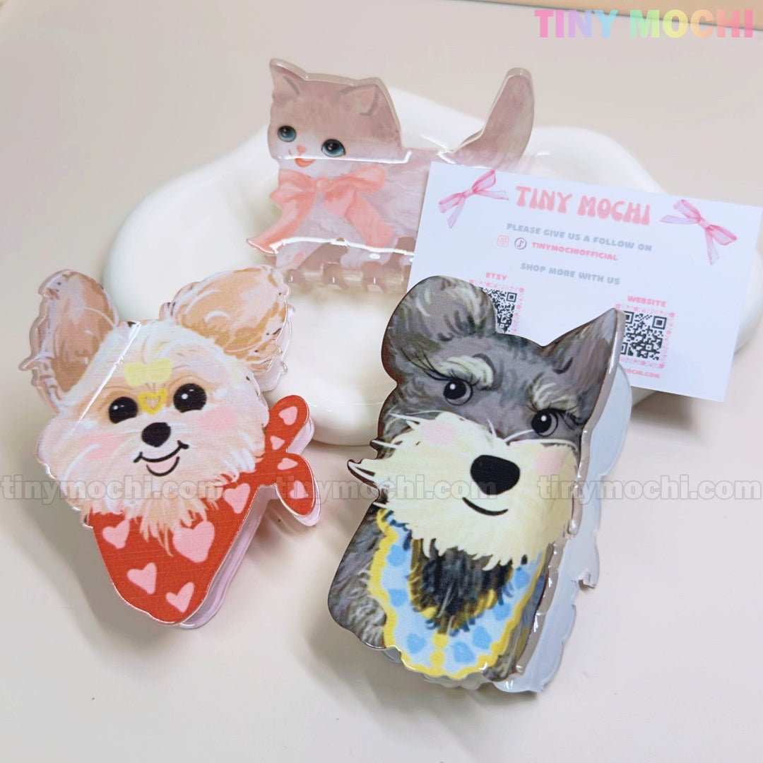 Three Kawaii Dog & Cat Hair Claw Clips from Tiny Mochi—featuring a cat, a dog with a red scarf, and one with a blue collar—are displayed on a white dish, with branded business card in the background.