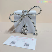 The Silver Teddy Bear Bow Keychain by Tiny Mochi is tied around a white box, with branded pastel card and QR codes in the background—making this cute bag charm accessory a perfect gift for women, teens, and kawaii lovers.