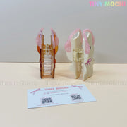 Two pastel hair claw clips—one shaped like a bunny and one like a bear, both wearing pink headphones—stand on a light surface next to a Tiny Mochi card. The perfect kawaii accessory for your aesthetic bag charm collection.