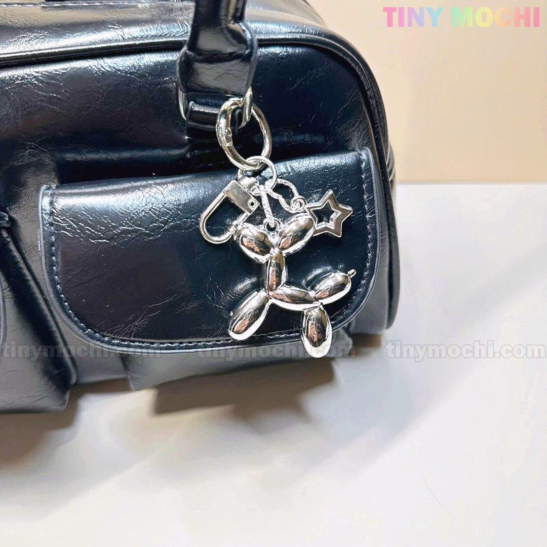 A shiny black bag with a Balloon Dog Keychain Silver Chrome Bag Charm with Star Accent by Tiny Mochi attached to the front pocket. Neutral background; Tiny Mochi appears in pastel colors in the top right corner.