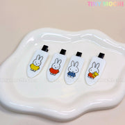 Four rectangular Tiny Mochi Kawaii Bunny Hair Clips for Girls, featuring cute cartoon bunnies in colorful outfits on resin alligator clips, are arranged on a wavy white dish. Black backs and vibrant illustrations decorate each barrette.