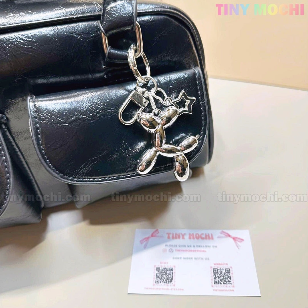 A Balloon Dog Keychain Silver Chrome Bag Charm with Star Accent by Tiny Mochi hangs from a black leather bag, while a branded Tiny Mochi card with QR codes rests on a beige surface, adding a playful metallic touch to your purse.
