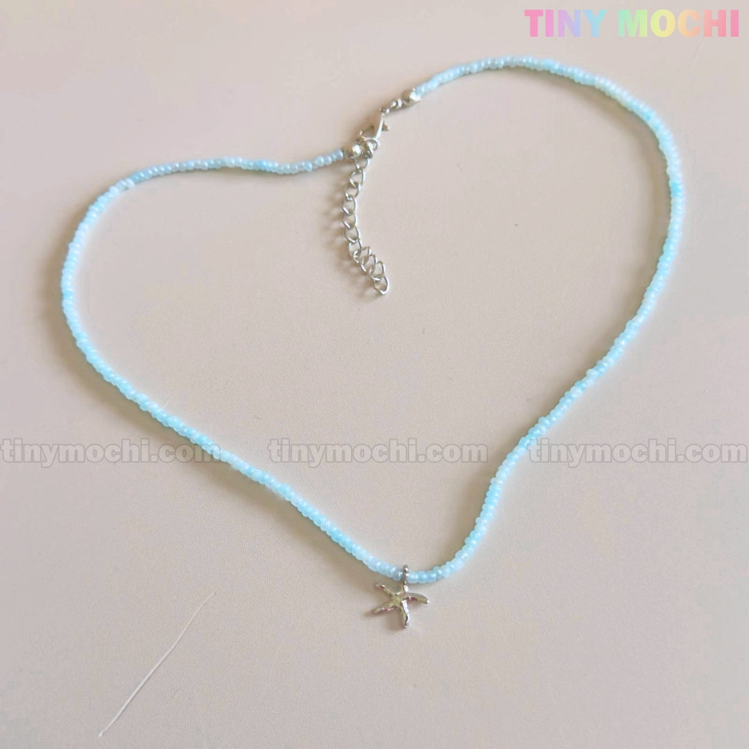 The Starfish Beach Necklace by Tiny Mochi features an iridescent blue pendant, arranged in a heart shape on a pale surface. TINY MOCHI appears in colorful letters at the top right, perfect for ocean or mermaidcore style.