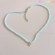 The Starfish Beach Necklace by Tiny Mochi features an iridescent blue pendant, arranged in a heart shape on a pale surface. TINY MOCHI appears in colorful letters at the top right, perfect for ocean or mermaidcore style.