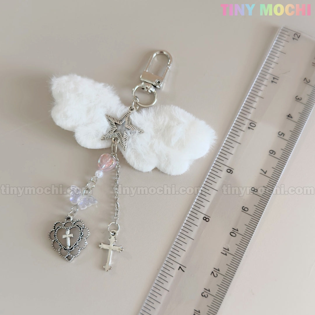 The Fluffy Angel Wing Keychain – Star & Cross Bag Charm Clip by Tiny Mochi features white plush wings with silver star, heart, cross charms, and beads. It sits by a clear ruler on a light surface. “Tiny Mochi” appears in colorful letters at the top right.