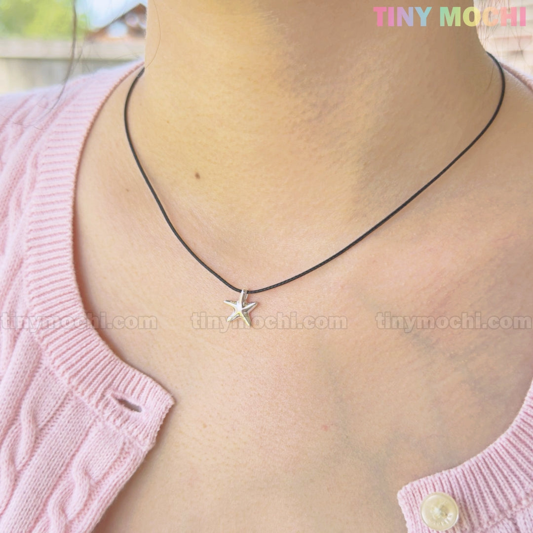 Outdoors, a person in a pink knit sweater wears the Tiny Mochi Sterling Silver Starfish Necklace 925 Charm on Black Cord—minimalist ocean jewelry and a handmade gift for him or her.