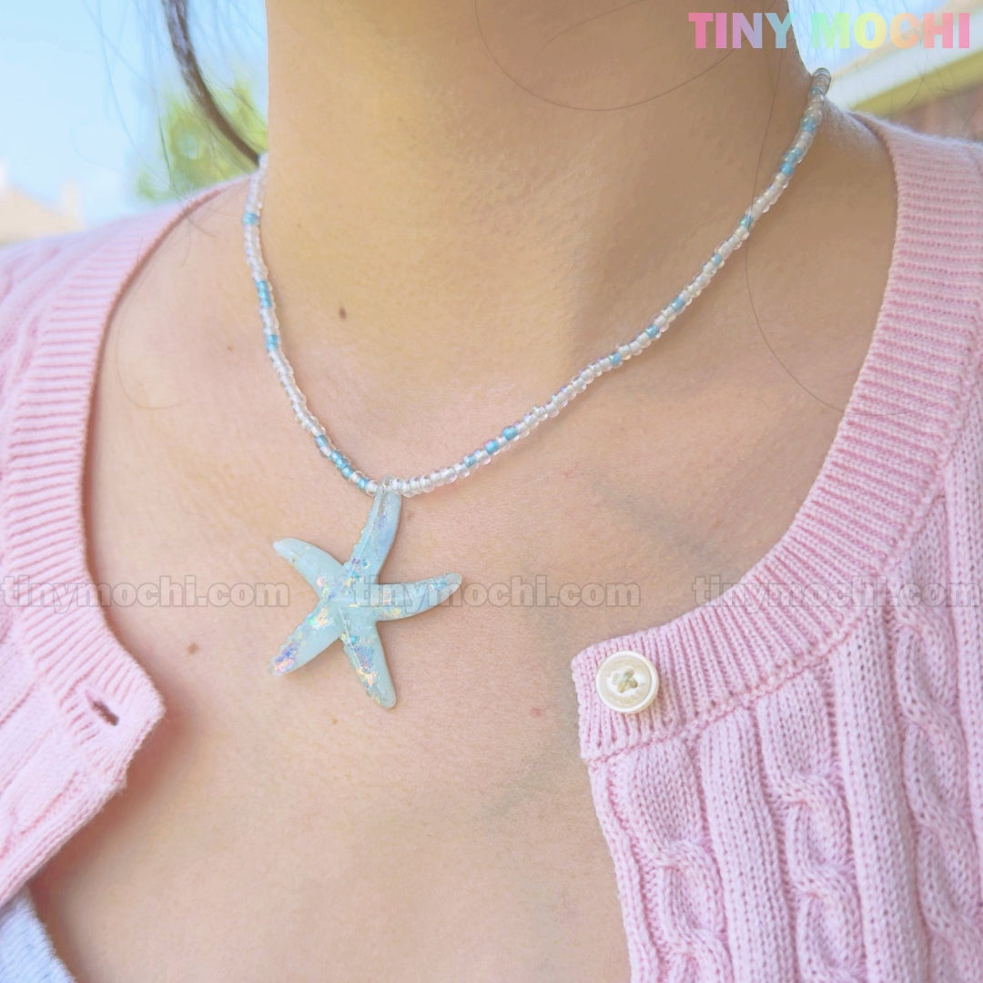 A person wears the Tiny Mochi Starfish Beach Necklace, featuring a handmade iridescent blue pendant choker, paired with a pink cardigan. The close-up highlights its ocean aesthetic. TINYMOCHI appears in the top right corner.